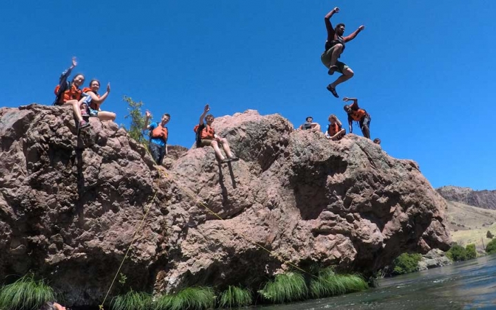 A student wearing safety gear jumps off a rocky ledge into a body of water. Others wearing safety gear sit on the ledge and watch.